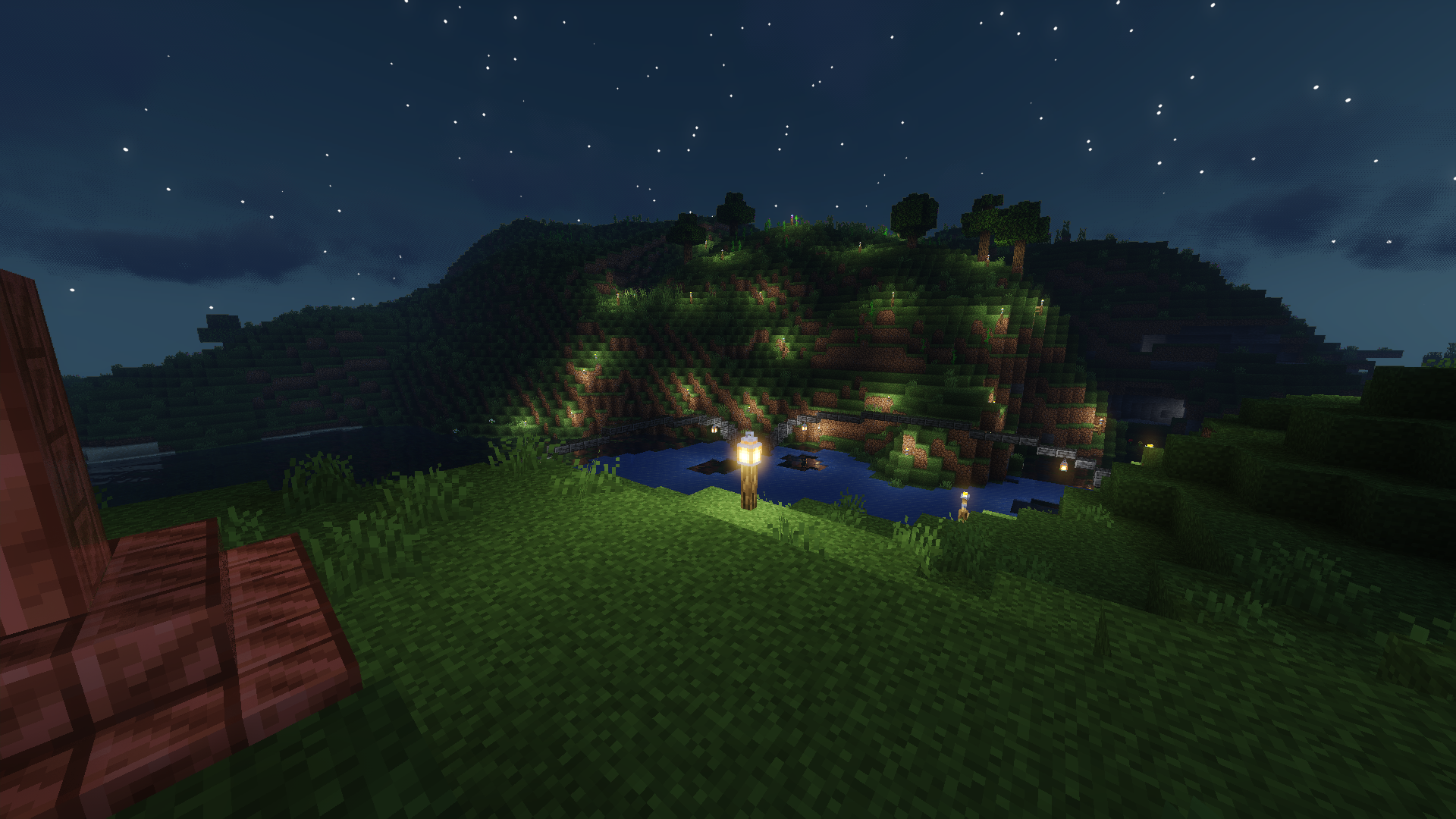 A screenshot of the game Minecraft. The screenshot shows a river at night, cutting through hills. Thin deepslate bridges with lanterns haning from beneath connect either side of the river. On both sides fence poles with lanterns on top are visible. Frosted Ice can be seen on the river due to the photographer forgetting they were wearing their Frost Walker boots.